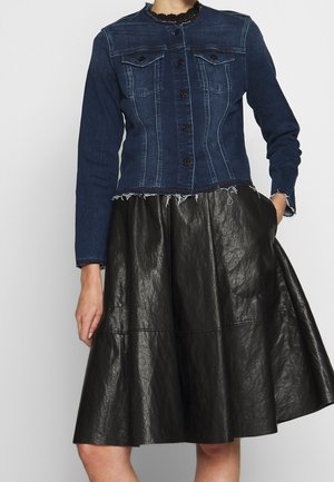 Woman wearing a dark blue denim jacket with frayed hem and buttons, paired with a black textured A-line skirt with pockets.