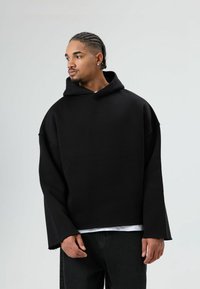 Zwarte oversized capuchontrui met wijde mouwen, gladde textuur en een casual pasvorm, gelaagd over een wit T-shirt, gecombineerd met zwarte broek.
