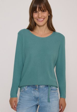 Femme souriante portant un pull en maille bleu sarcelle à manches longues avec un lien sur le côté et un jean bleu clair sur fond uni.
