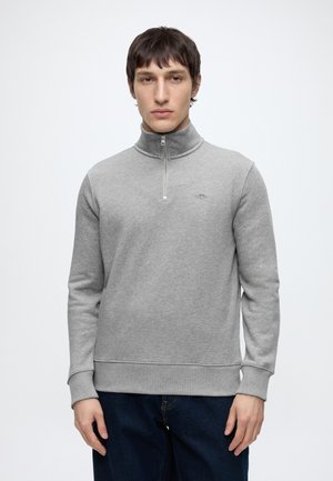 Jeune homme aux cheveux courts et foncés, portant un sweat-shirt gris clair à demi-fermeture éclair et un jean bleu foncé, debout devant un fond blanc uni.