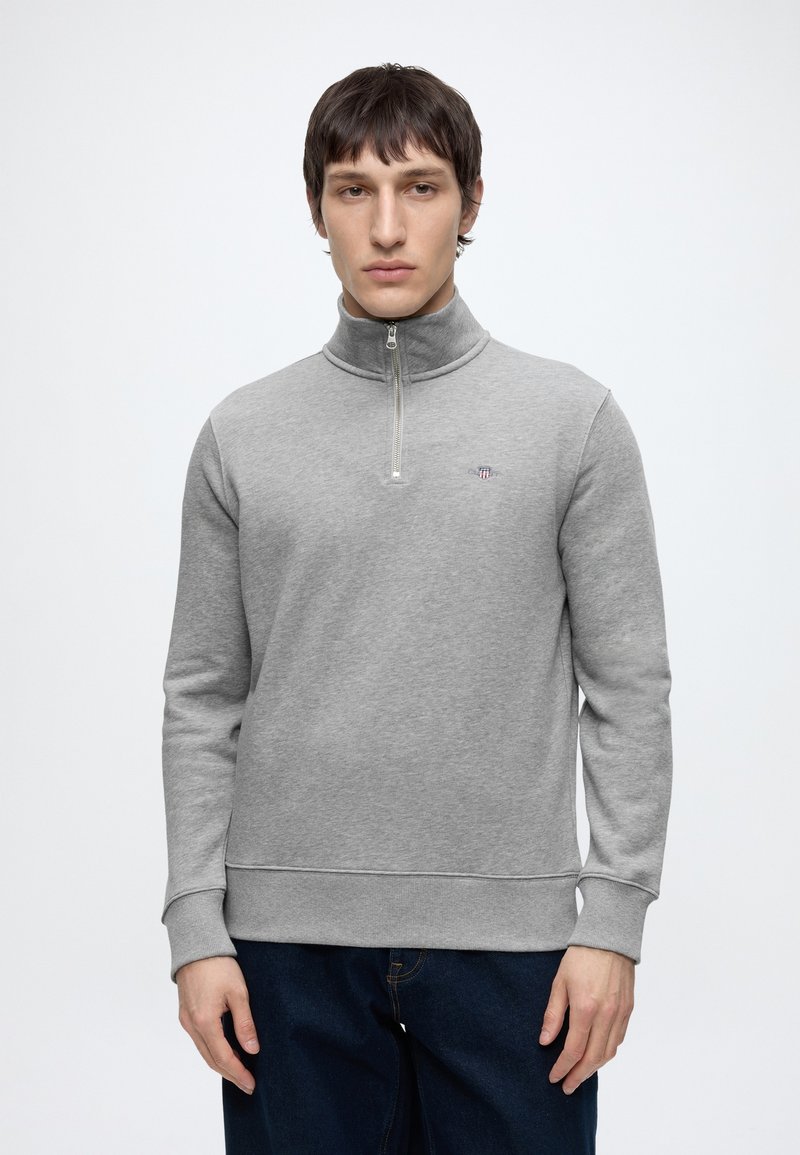 Junger Mann mit kurzen dunklen Haaren, der einen hellgrauen Halbzip-Sweatshirt und dunkelblaue Jeans trägt, steht vor einem schlichten weißen Hintergrund.