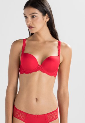 Reggiseno imbottito rosso con bordo in pizzo, dotato di spalline regolabili e un fiocco decorativo al centro. Tessuto liscio con accenti in pizzo floreale.
