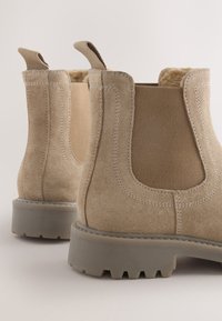 Beige ankelstøvler med ruskindslignende finish, elastiske sidepaneler og en kraftig grå gummisål med mønster.