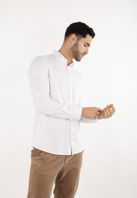 Hvit langermet skjorte med knappedekselkrage, kombinert med brune cordfløyelsbukser. Stoffet ser glatt ut med en tettsittende silhuett.