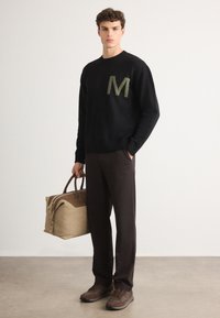 Suéter negro de punto con bordado verde de la "M", combinado con pantalones marrones y zapatillas marrones. El modelo sostiene un bolso de mano color beige con detalles marrones.