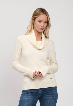 Lyshåret kvinde iført en cremefarvet sweater med snoet hals og blå jeans, stående mod en ensartet lys baggrund.
