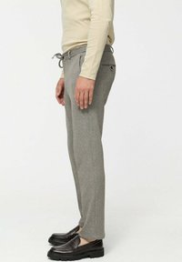 Man in een lichtbeige overhemd met lange mouwen, grijze broek met trekkoord en zwarte instapschoenen, staande in zijaanzicht op een witte achtergrond.