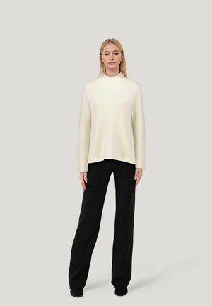 Cream ribbed pullover with a high neck and wide sleeves, paired with black wide-leg trousers and black footwear. Textured fabric, minimalist design.