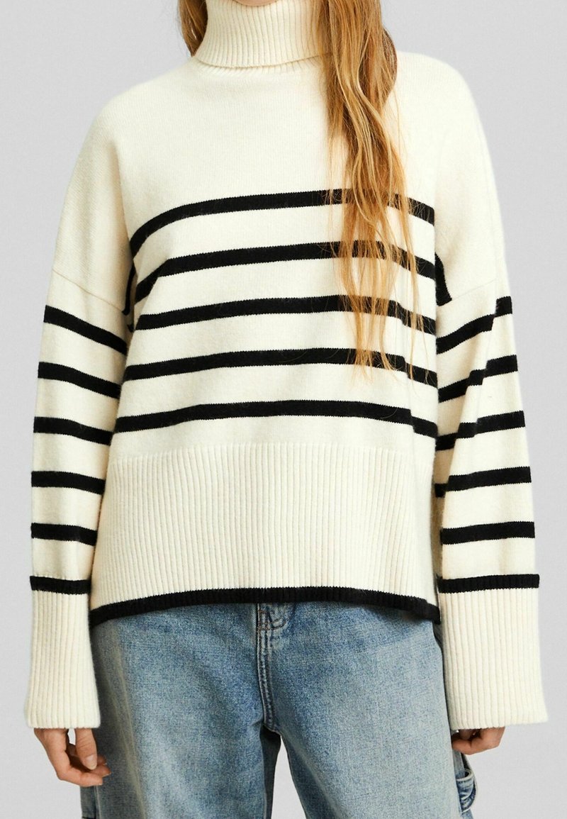 Cremefarbener Pullover mit schwarzen horizontalen Streifen, hohem Rippenkragen und weiten Ärmeln. Trägt dazu hellblaue, abgenutzte Jeans.