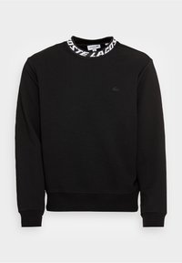 Sweat-shirt noir à manches longues avec poignets côtelés, col montant avec le texte "LACOSTE" et un logo crocodile brodé discret sur la poitrine.