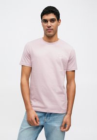 Απαλό ροζ βαμβακερό Τ-shirt με στρογγυλή λαιμόκοψη, κοντά μανίκια και ένα μικρό τετράγωνο λογότυπο στο στήθος. Το μοντέλο ποζάρει μπροστά σε απλό φόντο.