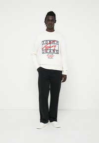 Weißer Strickpullover mit einem bunten "Tommy Jeans"-Logodesign. Kombiniert mit schwarzen Hosen und weißen Schuhen, steht vor einem einfarbigen Hintergrund.