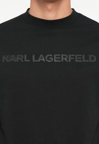 Črna majica z dolgimi rokavi iz mehkega bombaža, ki na sprednjem delu v poudarjenem sivo-natisnjenem besedilu "KARL LAGERFELD" v izraziti pisavi.