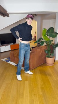 Henkilö mustassa poolopaidassa ja sinisissä farkuissa seisoo huoneessa, jossa on puulattia, ruskea sohva ja suuri vihreä kasvi ruukussa.