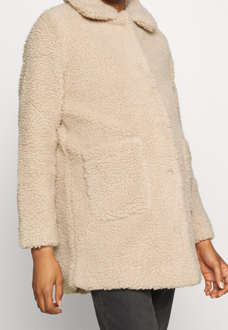 Veste en polaire sherpa couleur crème avec grande poche avant, col et manches longues, portée sur un pantalon foncé.