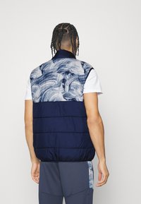 Puffa vesta s tmavě modrou spodní částí a vzorovanou světle modrou horní částí. Vybavená vysokým límcem a horizontálním prošívaným designem.