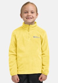 Glunderend kind met blond haar, gekleed in een gele fleece pullover met een kwart rits en zwarte broek, tegen een eenvoudige achtergrond.