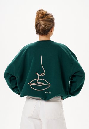 Person mit Dutt, die einen dunkelgrünen Pullover mit einer minimalistischen Gesichtslinienzeichnung auf dem Rücken und helle Hosen trägt.
