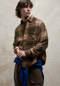 Rjava srajca s karirastim vzorcem in gumbi, z žepom na prsih, oblečena preko temno modre majice, modra jopa vezana okoli pasu in olivno zelene hlače s številnimi žepi.