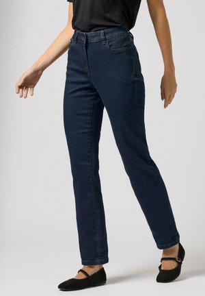 Person iført mørkeblå straight-leg jeans og sorte Mary Jane flade sko, stående med det ene ben let fremad mod en ensfarvet baggrund.