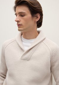 Hellbeiger gestrickter Pullover mit Schalkragen, der eine vertikale Rippentextur und eine halbe Knopfleiste aufweist. Über einem weißen Hemd getragen.