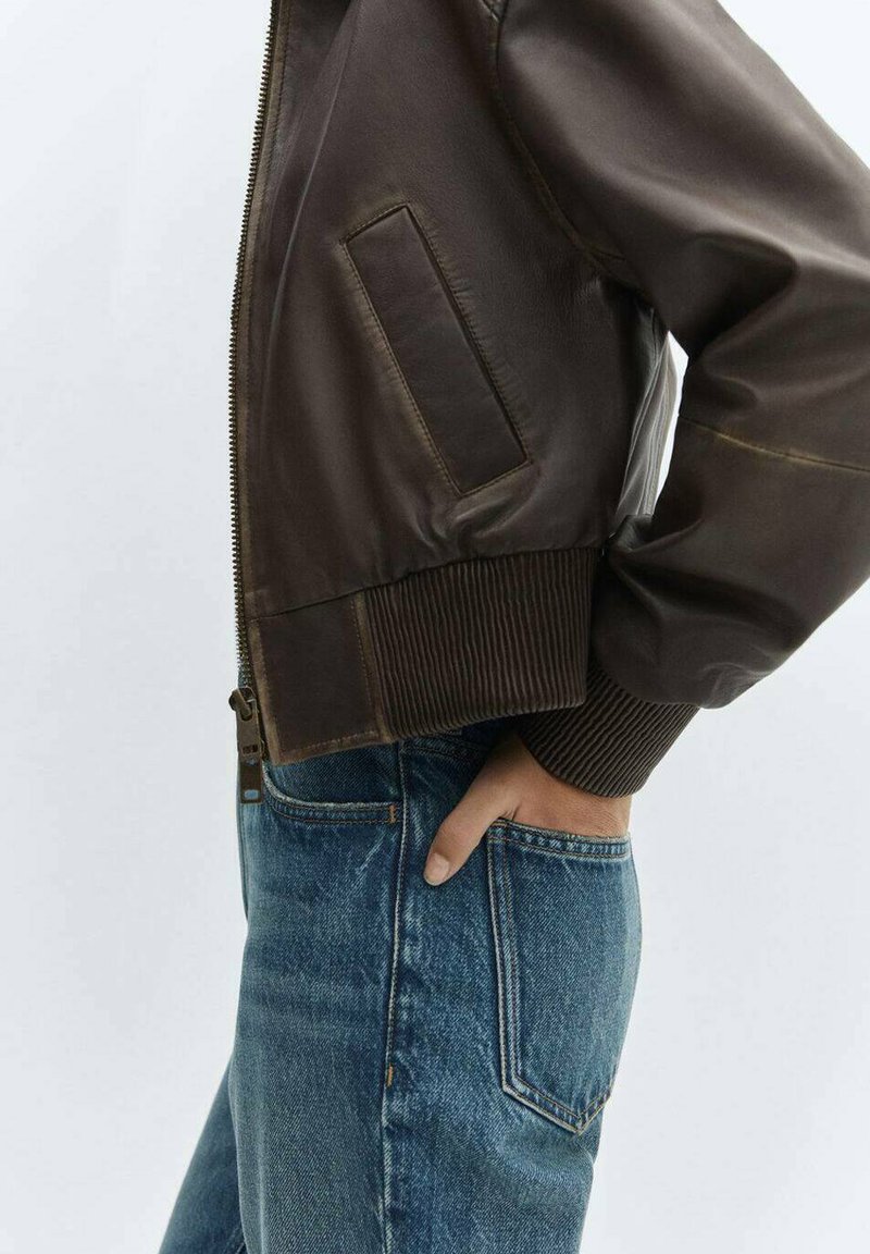 Braune Leder-Bomberjacke mit gerippten Bündchen und Saum, ausgestattet mit einer Seitenjacke. Kombiniert mit hochgeschnittenen blauen Jeans.