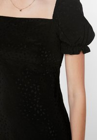 Musta mekko, jossa on neliömäinen kaula-aukko, puffihenkäyksiset hihat ja hienovarainen sävyjä vastaava tekstuuri. Mekossa on vartalonmyötäinen yläosa ja virtaava hame.