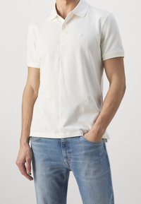 Kortärmad pique t-shirt i off-white bomull, med en spetsig krage, en enda bröstlogotyp och en rak fåll. Bärs med ljusblå jeans.