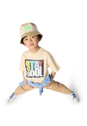 T-shirt beige avec le texte « STAY COOL » en lettres audacieuses et colorées. Chapeau bob vert clair, pantalon marron clair et baskets avec des lacets bleus.