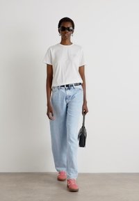 T-shirt bianco, jeans azzurri chiari, sneakers rosa, cintura nera, tiene una borsa nera, indossa occhiali da sole, con capelli corti e textured.