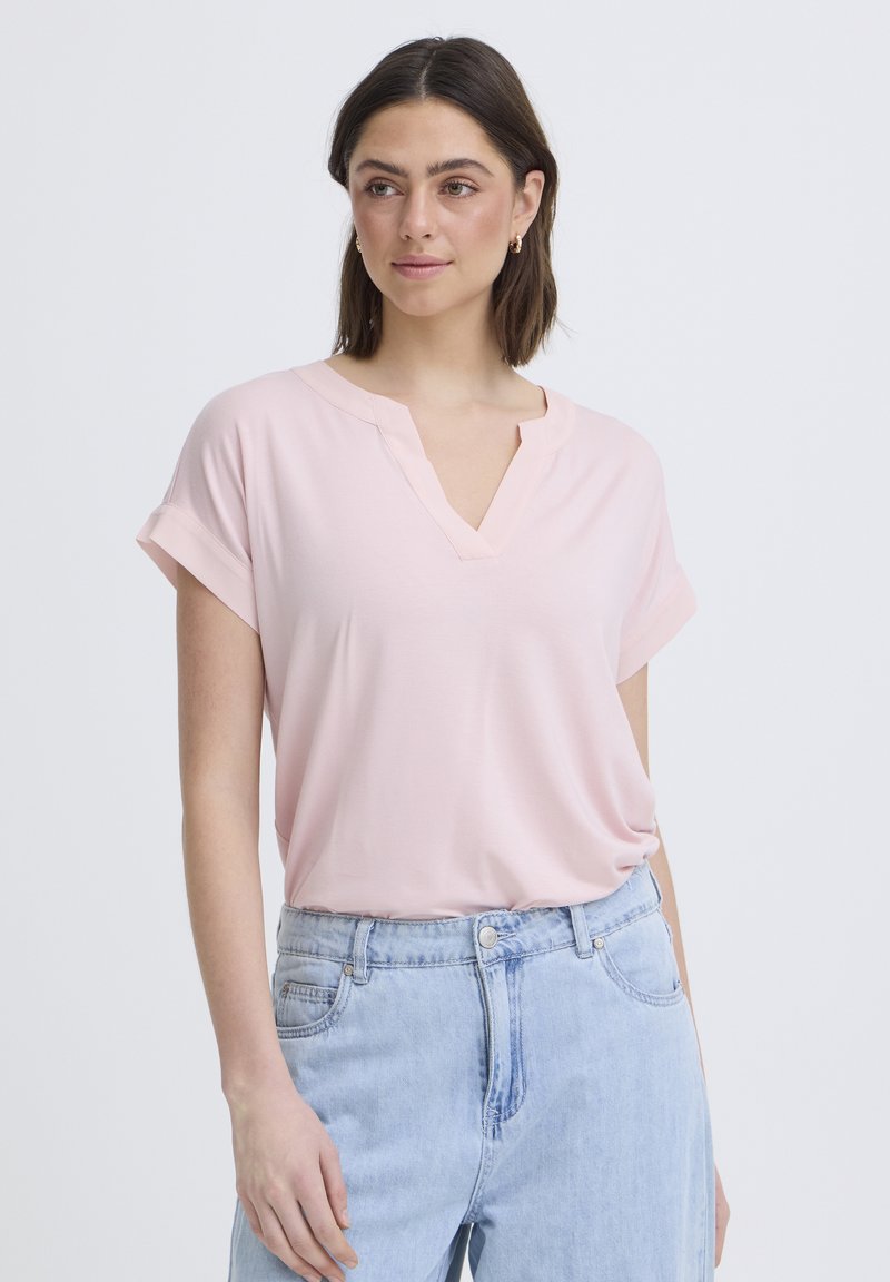 Femme portant un haut rose clair à manches courtes en V, rentré dans un jean taille haute bleu clair, debout devant un fond uni.