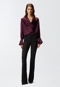 Bluza s kapuljačom u tamnocrvenoj boji s dugim rukavima i raširenim manžetama u kombinaciji s crnim krojenim hlačama, stvarajući elegantnu siluetu.