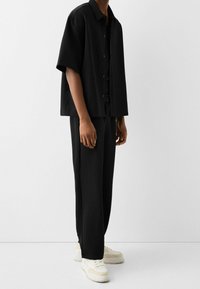 Person som står i svart kortärmad skjorta med knappar och svarta byxor kombinerat med vita och beiga sneakers framför en enkel ljus bakgrund.