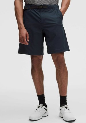 Mann trägt marineblaue Shorts mit Gürtel, schwarze Socken und weiße Sportschuhe, steht mit den Händen in den Taschen.
