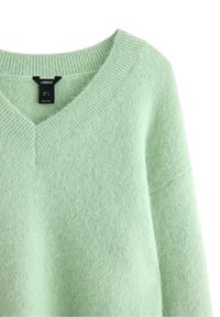 Harmaa vihreä neule, joka on valmistettu pehmeästä neulematerialista. Varustettu V-pääntiellä ja ribbaamalla reunalla. Tunnus näkyvissä, mukaan lukien koko tietoa.