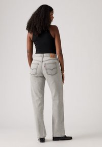 Žena s kovrčavom kosom, odjevena u crnu majicu bez rukava, svijetlosive traperice visokog struka i crne cipele, stoji protiv jednolične pozadine.