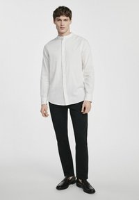 Biała koszula z kołnierzykiem stand, w połączeniu z czarnymi spodniami slim-fit i błyszczącymi czarnymi butami typu slip-on. Gładka tekstura materiału.