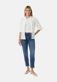 Frau steht da und trägt ein weißes Hemd über einem weißen Top, blue cropped Jeans und hellbeige Slingback-Sandalen vor einem schlichten weißen Hintergrund.