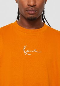 Tricou din bumbac portocaliu, cu guler rotund, având o semnătură brodată albă pe piept, textură netedă și o croială lejeră.