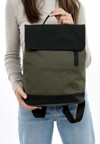 Olivgrüner Rucksack mit einer schwarzen Klappe, glatter Textur, verstellbaren Schultergurten und einem oberen Griff. Verfügt über ein Markenetikett an der Seite.