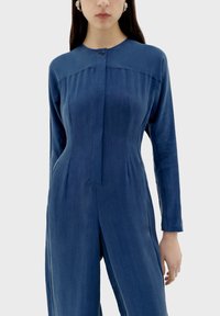 Denim jumpsuit met lange mouwen, ronde halslijn, sluiting met knopen aan de voorkant en zijzakken. Getextureerde stof in een diepe blauwe kleur.