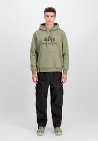 Mladý muž stojí, oblečený v olivovo zelenej mikine Alpha Industries, čiernych cargo nohaviciach a zelených maskáčových teniskách na pozadí bielej plochy.