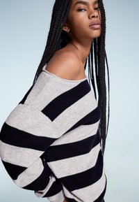 Fiatal nő hosszú fonott hajjal, vállra eső fekete és szürke csíkos pulóverben, egy világoskék háttér előtt pózol.