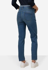 Tummansiniset farkut, joissa on slim fit -leikkuu, keskikorkea vyötärö, viisi taskua ja hieman lyhyempi pituus, yhdistettynä valkoisiin tennareihin.