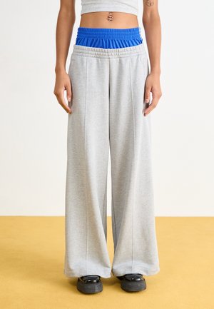 Kvinde iført brede grå joggingbukser over blå shorts, sorte chunky sko og en grå cropped top, der viser en navlepiercing og en tatoveret arm.