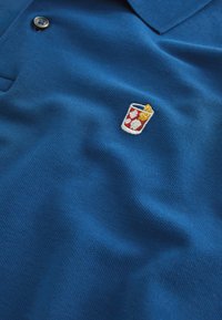 Polo bleu avec deux boutons et un petit verre à cocktail brodé avec des glaçons et une tranche de citron sur la poitrine.