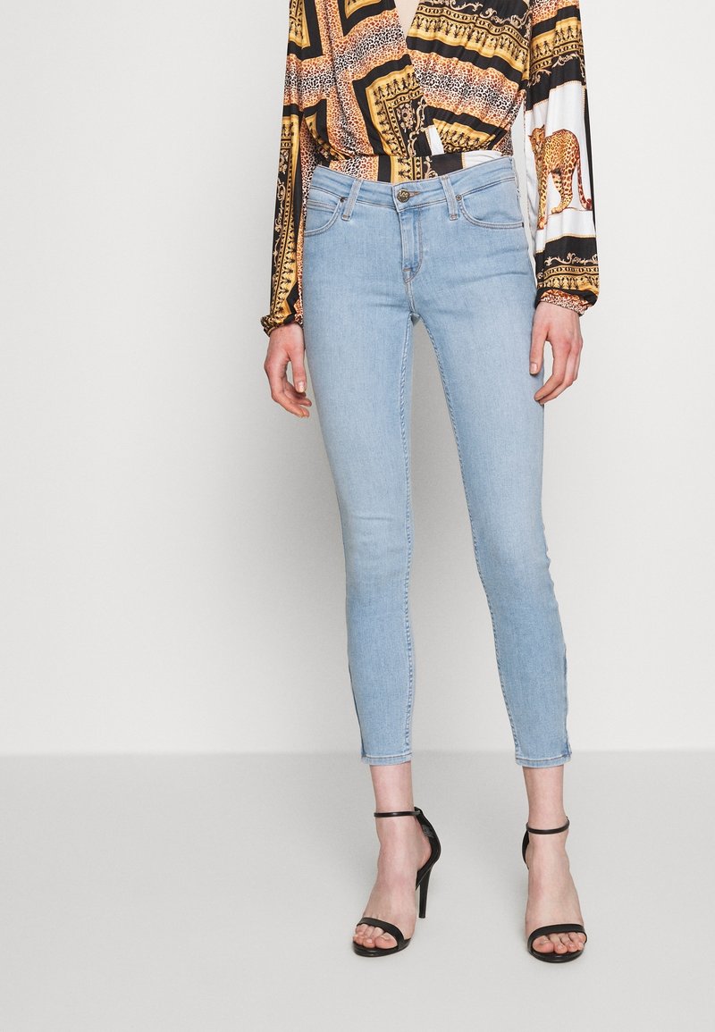 Jeans skinny azul claro de mezclilla, con cintura de media altura, diseño de cinco bolsillos y longitud recortada, combinados con sandalias de tiras negras.