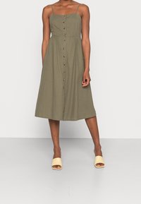Robe midi vert olive avec des bretelles fines, un devant à boutons et une taille froncée. Le modèle l'a associée à des mules jaunes à bout ouvert.