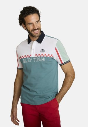 Homme souriant portant un polo bleu, blanc et menthe à carreaux rouges et un pantalon rouge, debout avec une main dans la poche.