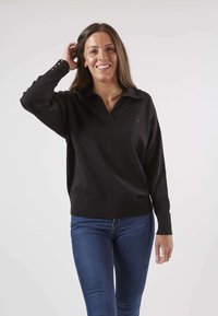 Pull noir en tricot avec un col en V et des manches longues. Présente une coupe décontractée et un ourlet côtelé. Porté avec un jean bleu ajusté.